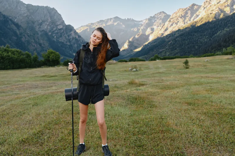 A focused female photographer stands in a scenic meadow surrounded by distant mountains. She holds a camera with a strap, ready to shoot, exuding calm concentration and a spirit of outdoor adventure.