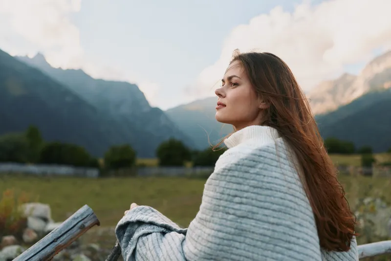 A young woman sits outdoors wrapped in a light gray blanket, gazing into the distance with rugged mountains in the background.