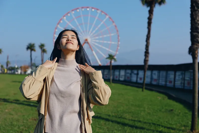 Woman outdoors under bright sunlight, lifting her jacket with a serene smile near a park and a ferris wheel, capturing carefree leisure and warm mood in an open coastal setting for travel mood now