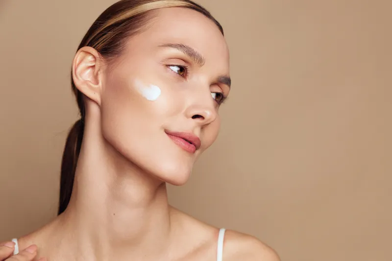 Young woman with clean skin applying cream on cheek, natural makeup, smooth complexion, soft lighting, neutral background, beauty skincare concept