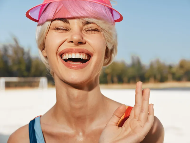 Joyful closeup of a smiling woman wearing a pink visor, sunlit beach scene in the background, vivid colors, and carefree summer happiness captured in bright light.