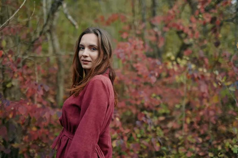 Authentic portrait of a woman in a wine red coat standing among autumn foliage, conveying genuine emotion, quiet confidence, and credible mood under soft daylight.