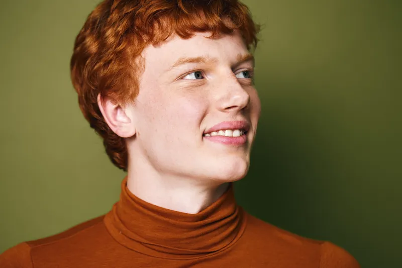 Young man with red hair wearing brown turtleneck looking sideways with smile against green background in studio. Closeup portrait of positive person with freckles and casual outfit.