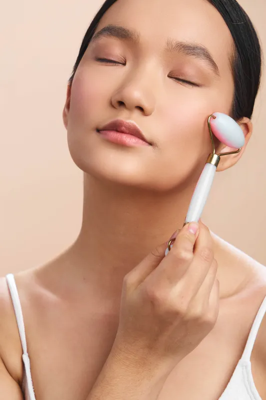 Asian woman enjoys a tranquil skincare moment, using a facial roller on her cheek with eyes closed, in a soft neutral setting that conveys calm, focus, and a gentle wellness routine.