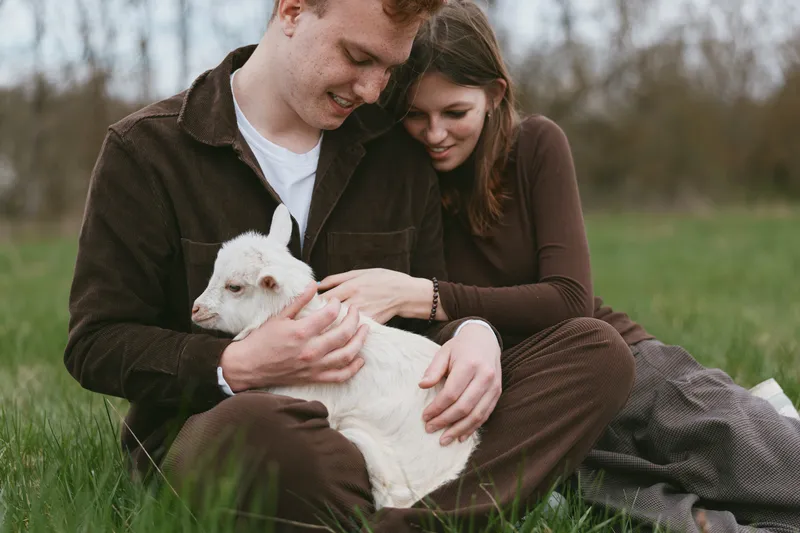 Couple sits on the grass outdoors, softly embracing a white goat kid, sharing a warm moment of affection in a rural park, casual clothing, calm natural setting.