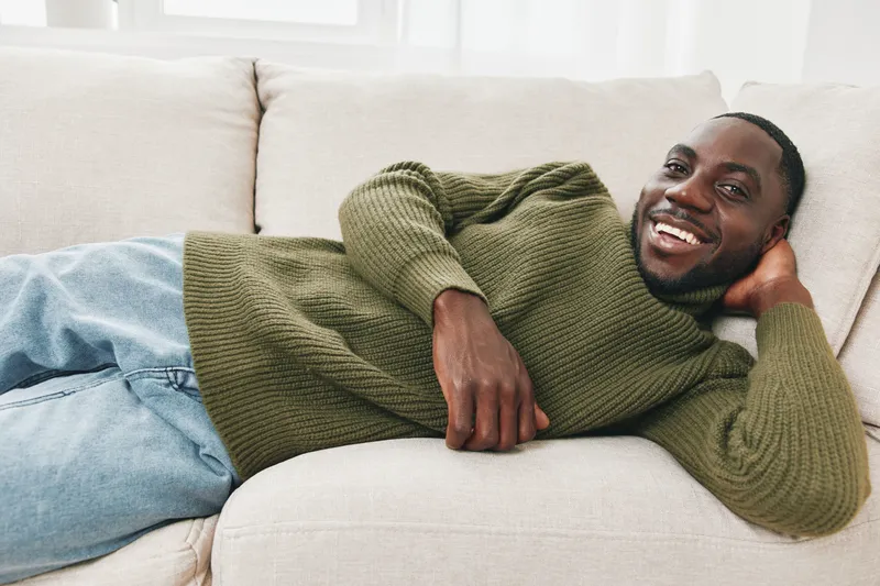 Smiling Black man relaxing on a cozy sofa in stylish green sweater, showing happiness and comfort in a bright, modern living room
