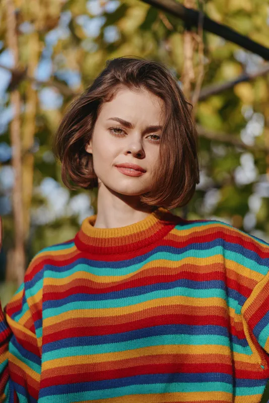 Woman portrait of a young adult in her 20s wearing a colorful striped sweater, smiling softly with bob haircut outdoors among autumn trees, casual lifestyle fashion shot.
