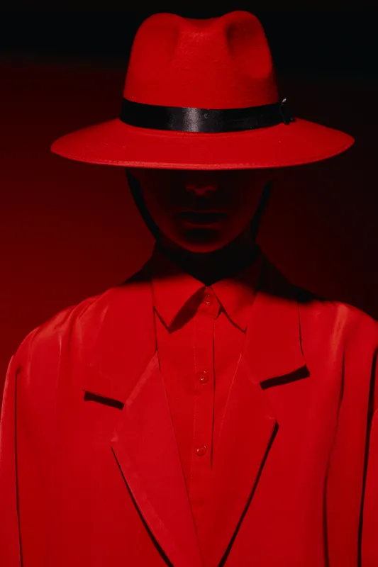Fashion creative woman wears wide brim red hat and matching red suit in low key lighting. Elegant style with vivid color emphasis and refined details in strong shadows. Beauty concept.
