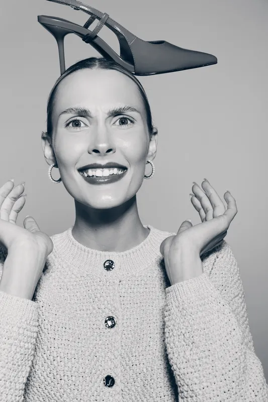 White caucasian woman with light skin smiles widely holding a high heel shoe on her head. Studio portrait with fashion style and playful concept in black and white photography.