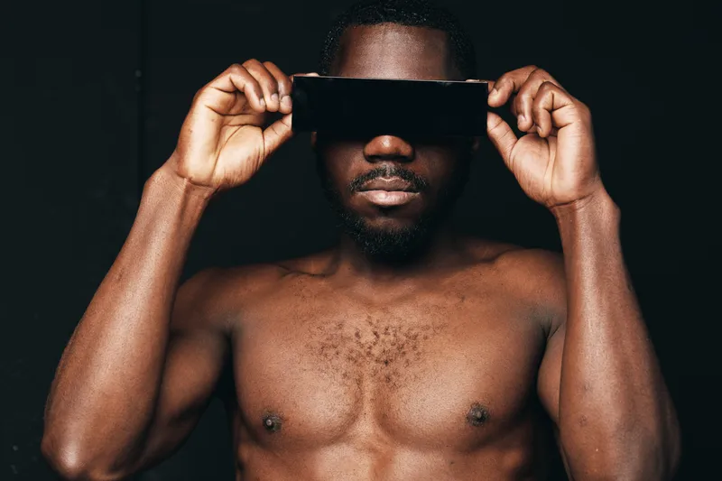 African man with dark skin and muscular body holding black glasses in creative conceptual visual style. Portrait of shirtless male with confident expression and artistic lighting on black background.