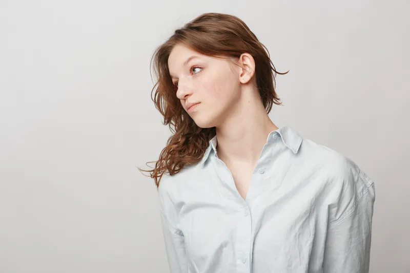 Young woman with curly brown hair wearing a white shirt looks sideways against a plain light background. Concept of natural beauty, casual style, and thoughtful mood in studio.