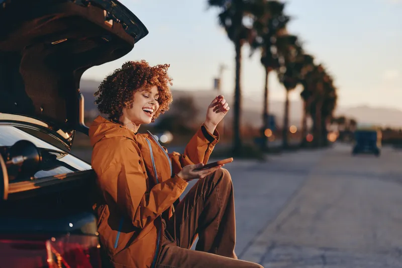 Woman lifestyle outdoors, smiling with curly hair, beside a car with open door at sunset, wearing a jacket, travel ready, warm sunlight, soft city street background