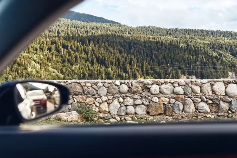 car window mirror reflecting road, stone wall and mountain forest landscape, authenticity inclusivity