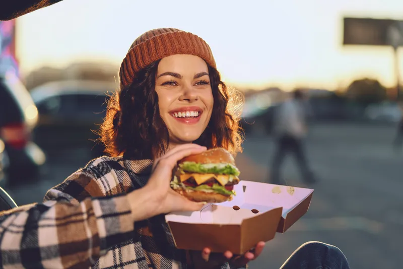 Woman burger smile outdoor beanie takeout candid golden authenticity in a mindful living scene; emotional storytelling captures a joyful urban portrait at warm sunset with genuine candid lifestyle.