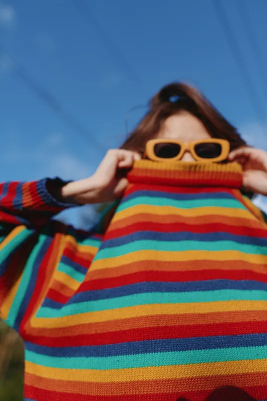 Striped sweater young woman sunglasses outdoor colorful covering face casual outfit blue sky, mid twenties model pulling turtleneck up, playful mood, cozy knitwear portrait for lifestyle and fashion.