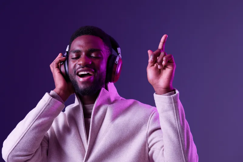 Young African man enjoying music with headphones, dancing against a vibrant purple background, showcasing joy and relaxation in winter fashion