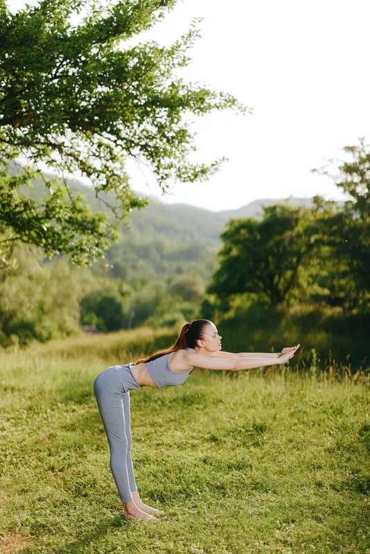 Young woman practicing yoga outdoors in a serene natural setting, wearing a gray athletic outfit, expressing calmness and focus, surrounded by greenery and warm sunlight