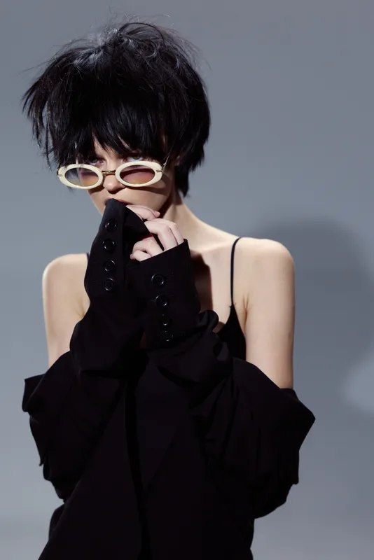 Stylish woman with short hair wearing white sunglasses and black outfit posing against gray background, fashion, modern, edgy, portrait, contemporary style