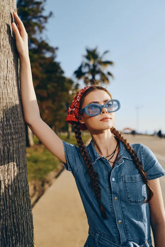 young woman in denim dress and blue sunglasses leaning on palm tree outdoors on sunny day with red bandana and braided hair