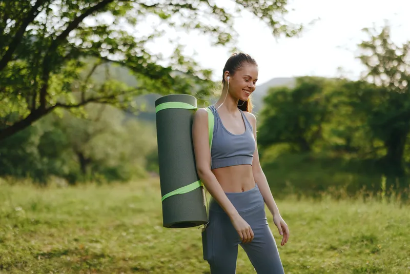Young woman in activewear with yoga mat enjoying nature, embodying wellness and fitness, surrounded by greenery in a serene outdoor setting