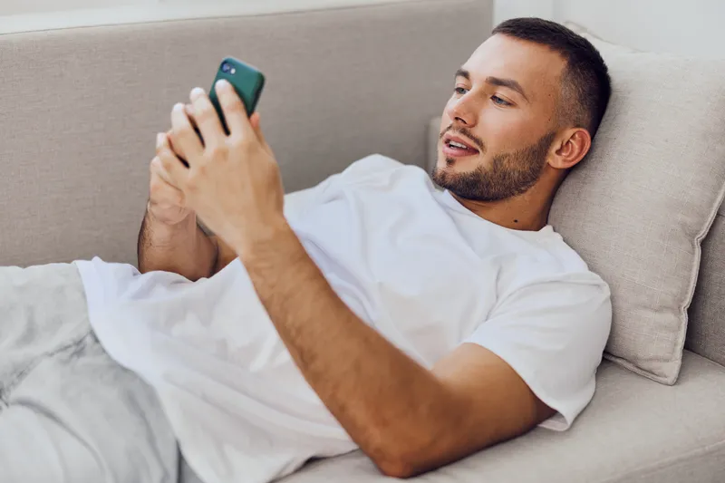 Relaxed man using smartphone while lounging on a couch, showcasing modern lifestyle and technology trends of 2025.