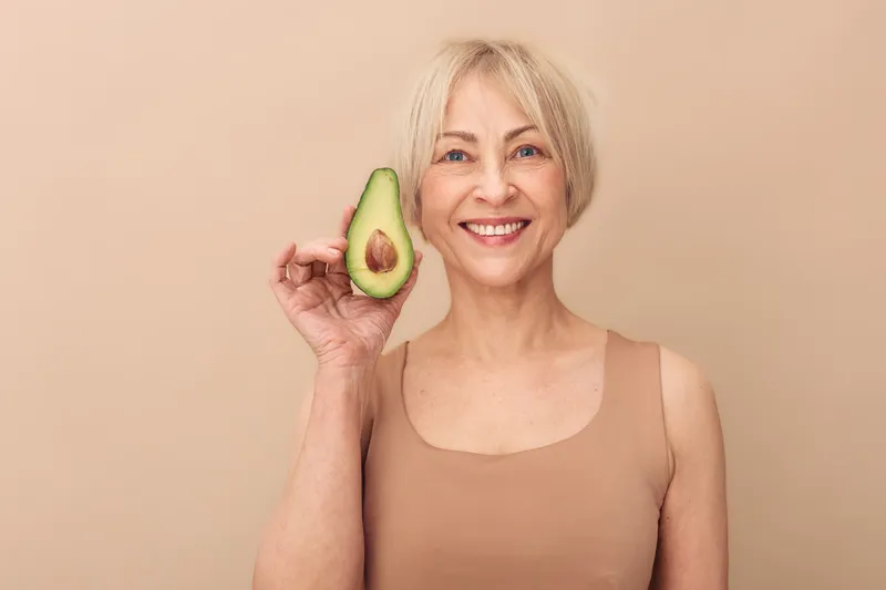 Smiling mature woman with short blonde hair holds half of fresh avocado near face on beige background. Concept of healthy lifestyle, nutrition, wellness, and natural beauty for senior women.
