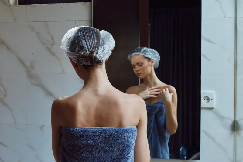 woman in shower cap wrapped in towel standing in modern bathroom facing mirror, skincare routine, personal care, wellness, calm atmosphere