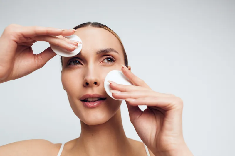 Young woman with clear skin applies cotton pads to her face in a clean studio, performing a calm skincare routine under soft warm lighting.