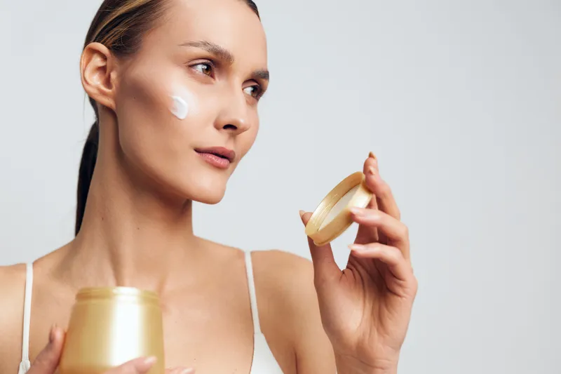 A young woman applies face cream on her cheek while holding an open jar and lid in a bright studio. Concept for skincare, beauty routine, hydration, and self care at home.
