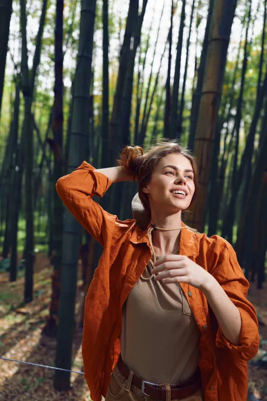 Woman in bamboo forest portrait smiling, wearing orange jacket and ribbed sweater, outdoors among tall green stalks. Happy young traveler enjoying nature, light and peaceful woodland mood.