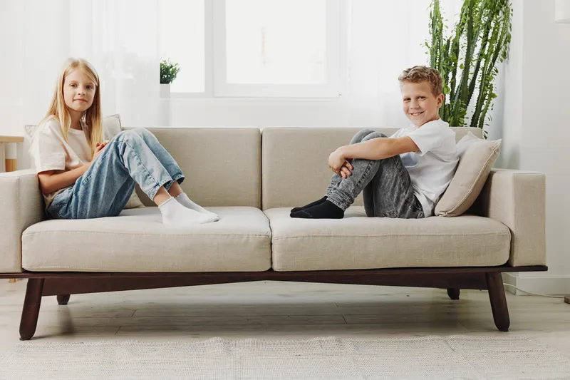 Two children sitting apart on a modern beige sofa in a bright living room, each looking at the camera with relaxed expressions. Concept of distance, family, and home life.
