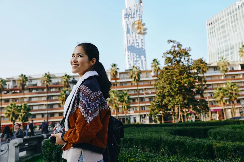 Traveler, backpack, woman in a warm jacket walking through a sunny urban park with palm trees and modern buildings in the background, capturing a cheerful daytime stroll