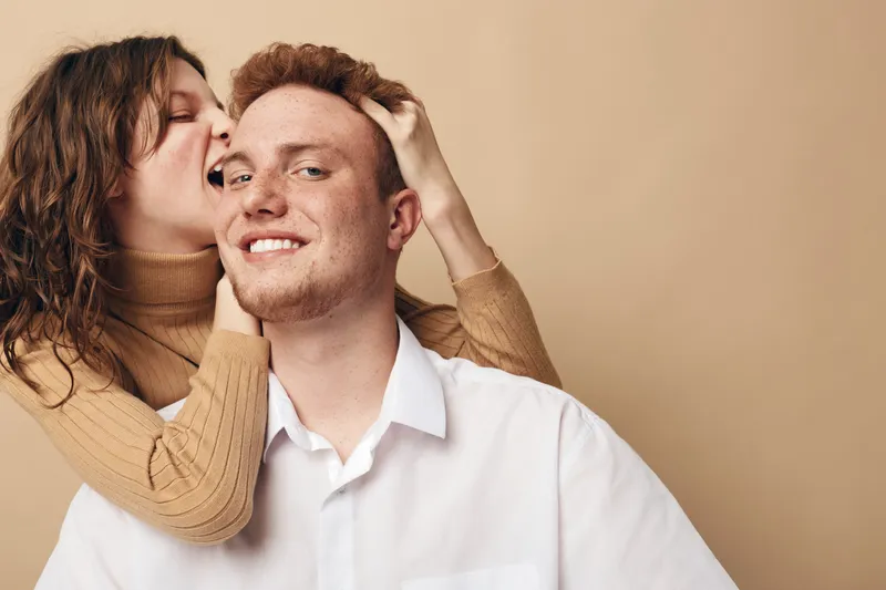 Happy young couple in casual clothes share playful moment, woman gently bites man's head. Warm beige background creates cozy and intimate atmosphere of love and fun.