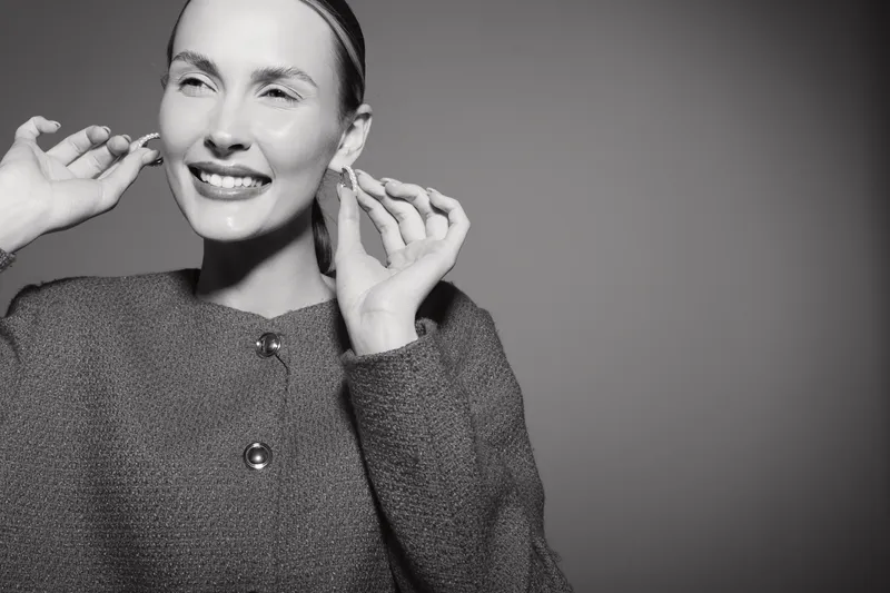 Happy Caucasian woman with light skin and sleek hair smiles broadly adjusting earrings in studio. Concept of joy, fashion, accessories, and beauty portrait in black and white.