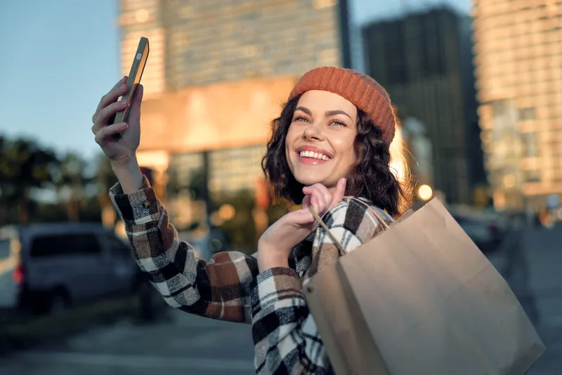 Woman taking selfie with shopping bag, smile and candid authenticity in urban sunset. Golden hour glow highlights joyful portrait, mindful living and emotional storytelling in city lifestyle.