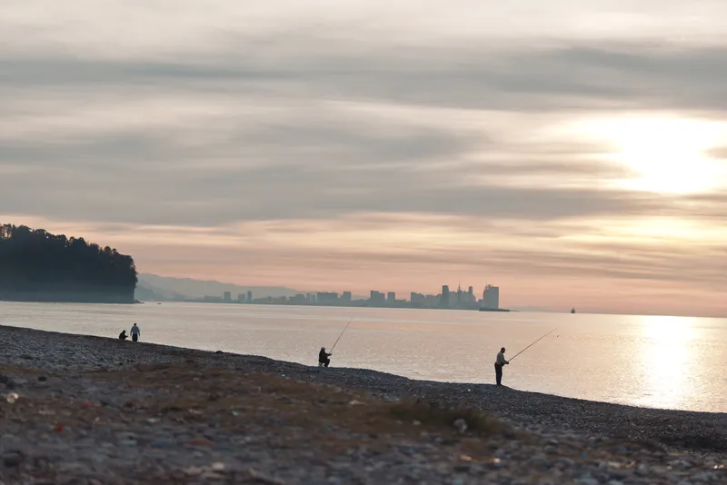Beach at sunset with calm sea and distant city skyline, fishermen along the shore, silhouettes moving against warm sky, reflective water and tranquil coastal scene