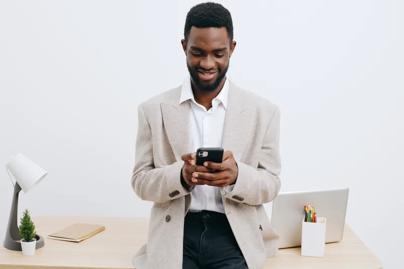 Smiling man with dark skin using smartphone in modern office setting, casual clothing, standing near desk with laptop, stationery, and potted plant, isolated on plain white background, studio shot, pe