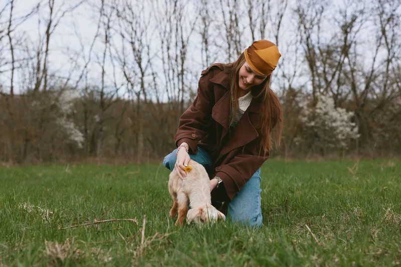 Smiling woman in a brown coat guides a playful goat kid across a grassy field, capturing a warm countryside moment. Lifestyle, nature, and animal bonding shine through soft outdoor light.