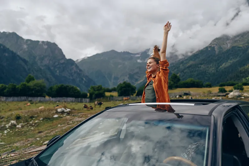 Adventure traveler raises arms from a sunroof as a car sits in a dramatic mountain landscape, conveying freedom, joy, and the thrill of an open road.