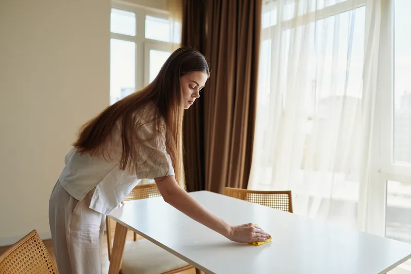 Woman cleaning table in bright home dining room, wiping surface with yellow cloth by window. Domestic chores, tidy interior, natural sunlight, focused household routine and care.