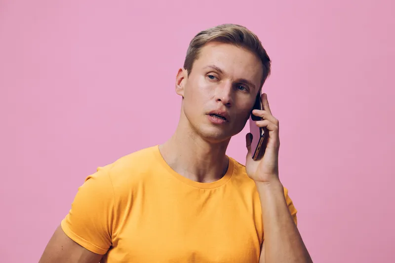 Close-up of young man with short brown hair and fair skin wearing a yellow t-shirt talking on smartphone against pink background, casual style, positive mood, studio shot. People lifestyle concept