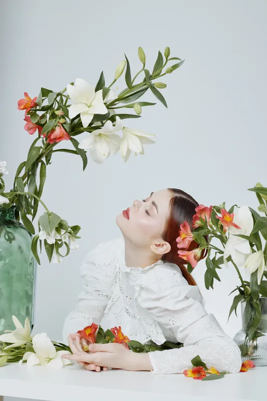 A woman in a white blouse surrounded by vibrant flowers, exhibiting a serene expression, perfect for concepts of beauty and nature appreciation