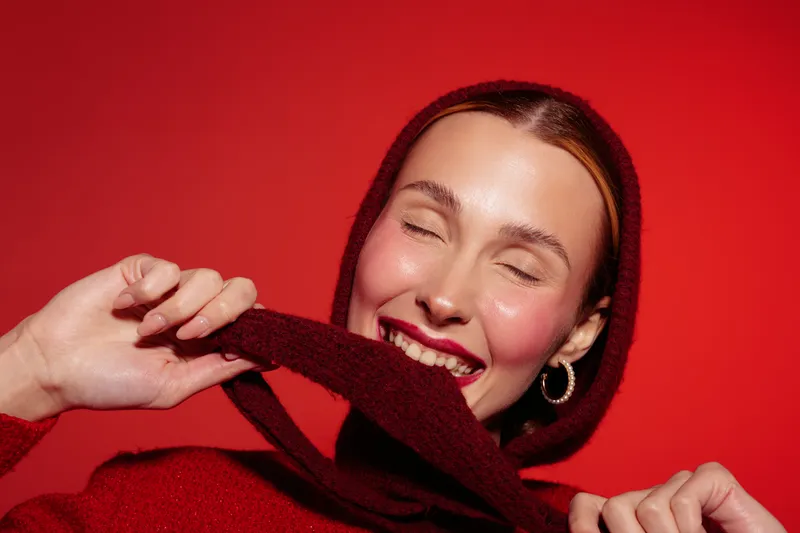 Smiling woman in red sweater and hood enjoys playful moment with eyes closed on red background. Concept of happiness, warmth, fashion, and cozy winter style with joyful expression.