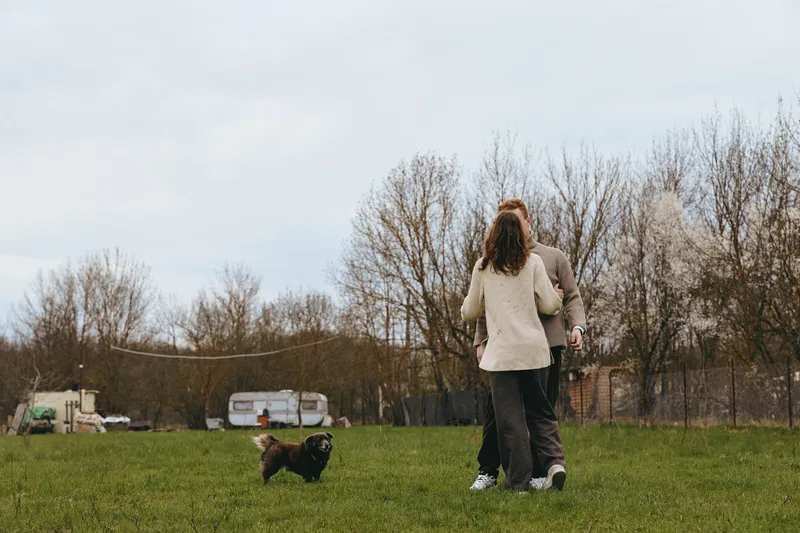 A couple walks a dog across a grassy field on a cool afternoon, casual outfits, bright sky, serene outdoor scene. A lifestyle moment showing companionship, exercise, and everyday routine.