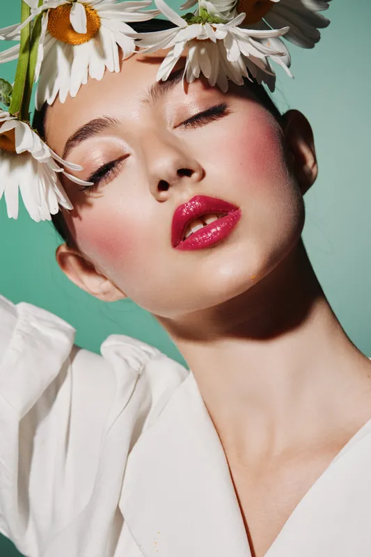 Portrait of a young woman with glossy red lipstick and lush blush wearing white blouse and daisy crown on mint background. Concept creative beauty, fashion, makeup artistry, elegance.