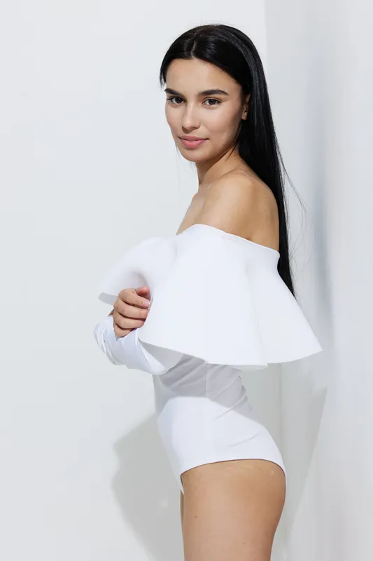 woman sport fitness portrait with long dark hair, wearing white off-shoulder fitness outfit, standing against a light wall, natural confident expression