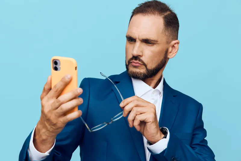 A focused business professional man on a vivid blue background, examining his smartphone with a furrowed brow and confident posture.