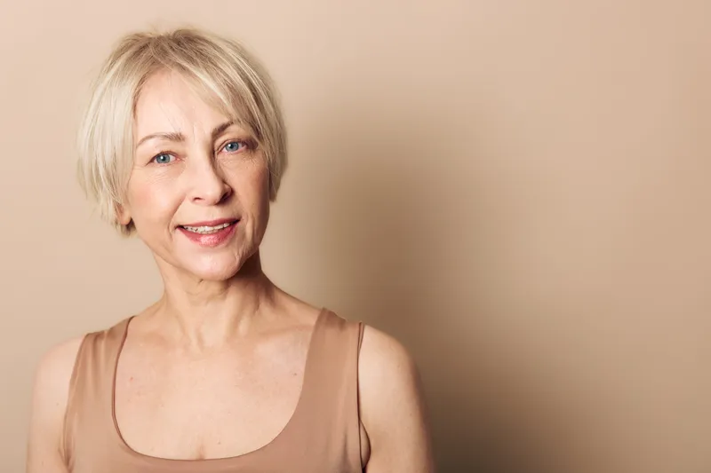 Smiling middle aged woman with short blonde hair wearing beige tank top stands against light brown background. Natural beauty portrait with soft lighting and warm neutral tones for wellness concept.