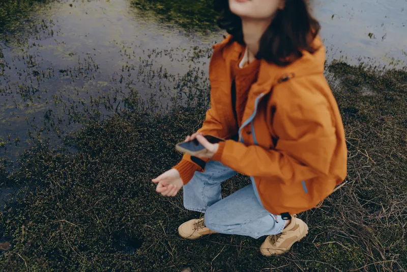 Woman lifestyle outdoor autumn scene, orange jacket and jeans, kneeling on street pavement, close up candid portrait. Soft natural light, casual fashion, calm mood with hands relaxed.