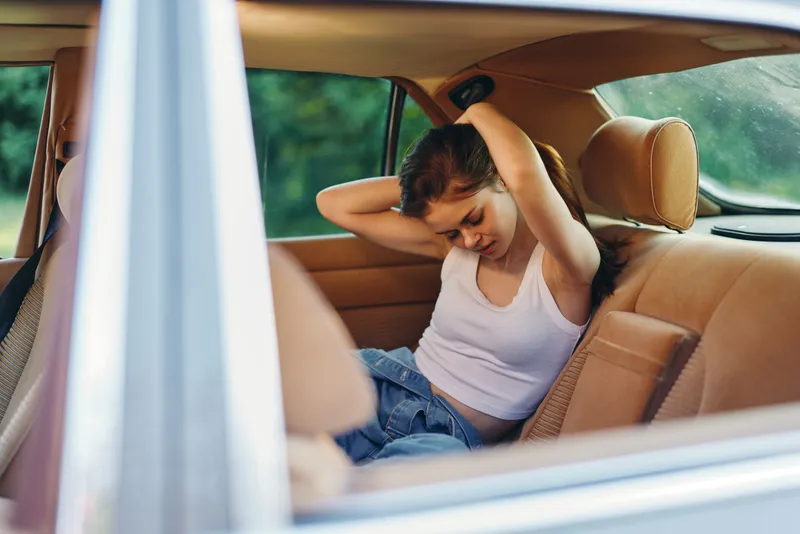 A thoughtful young woman in a white tank top sitting in the backseat of a vintage car, lost in deep contemplation against a soft green background.
